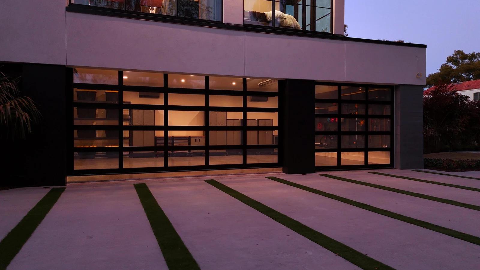 Two bays of full view glass garage doors glowing warm at dusk over a grass strip driveway