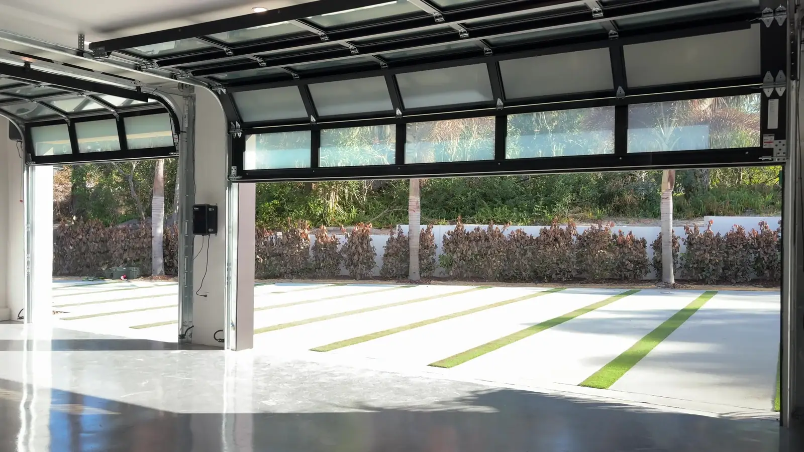 Looking out of a garage through an open full view glass door onto a sunlit driveway