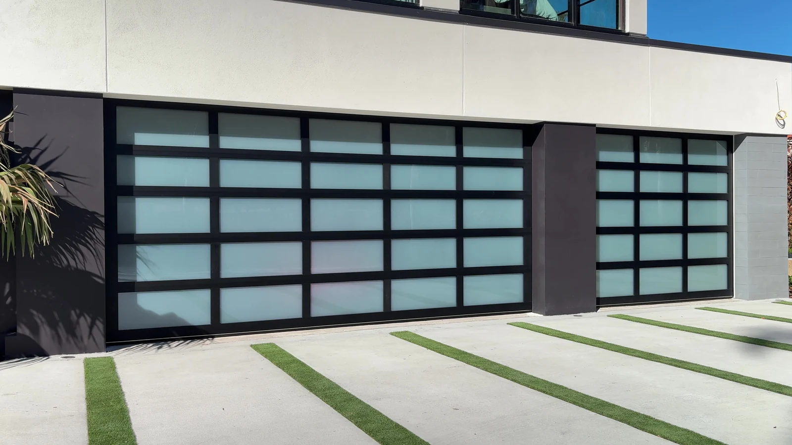 Black framed frosted glass Clopay Avante doors on a modern white stucco home, with a palm and a grass strip driveway