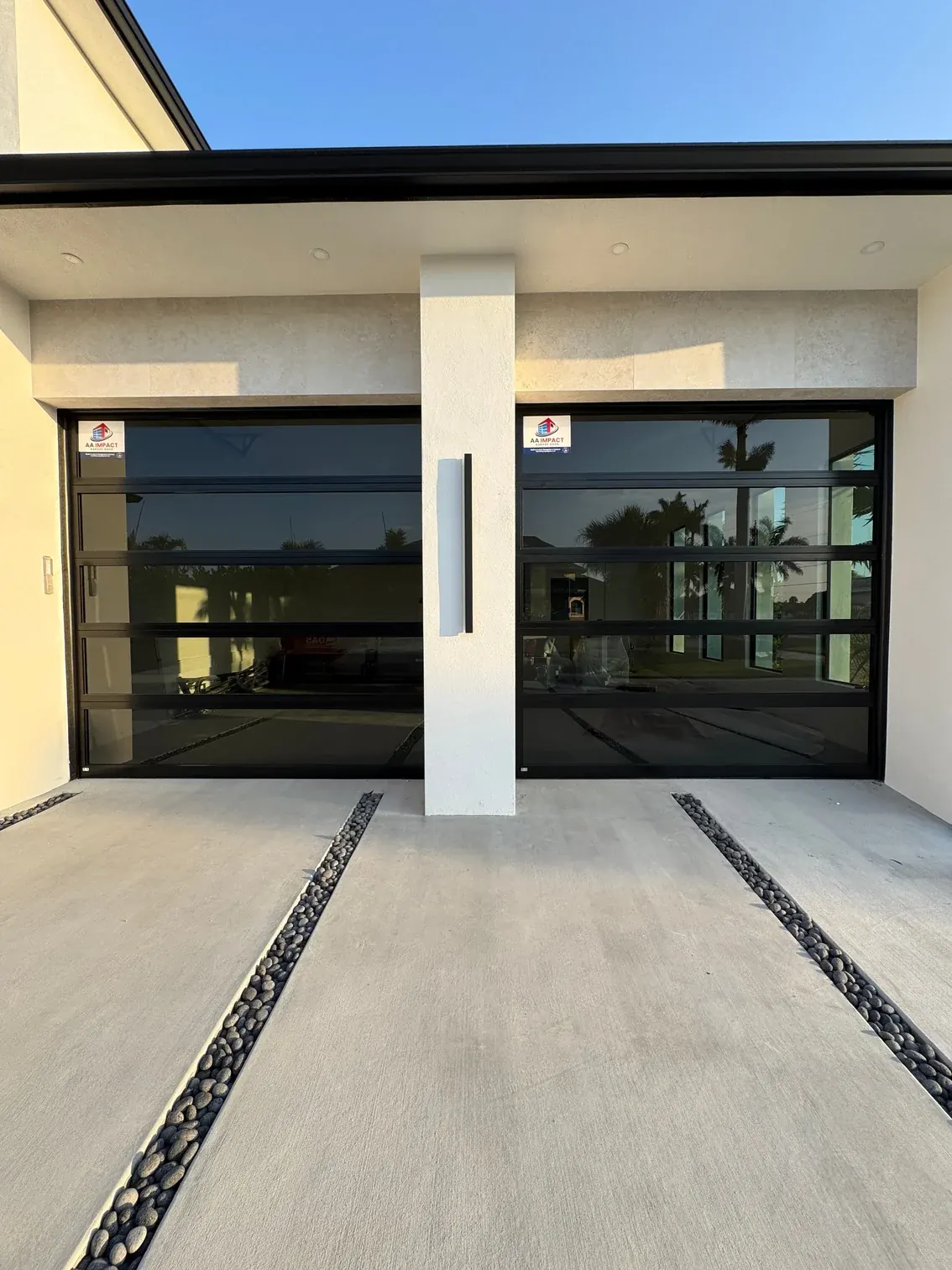 Two AA Impact Series 1200 single lite impact glass garage doors, black frames and dark glass on a white home