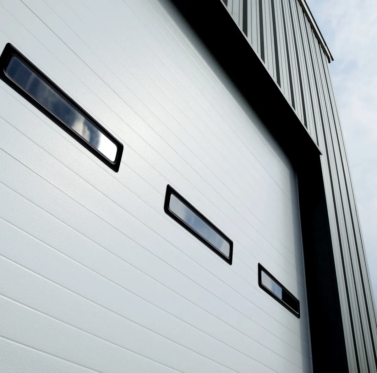Amarr 2432 full view commercial door, low angle of a white pencil groove door with three slim black framed windows on a metal building. Cover of the 2432/2732 b