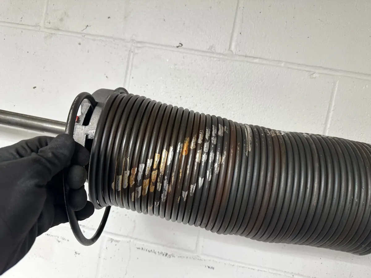 A gloved hand holding a broken torsion spring, with the gap where the coil snapped clearly visible