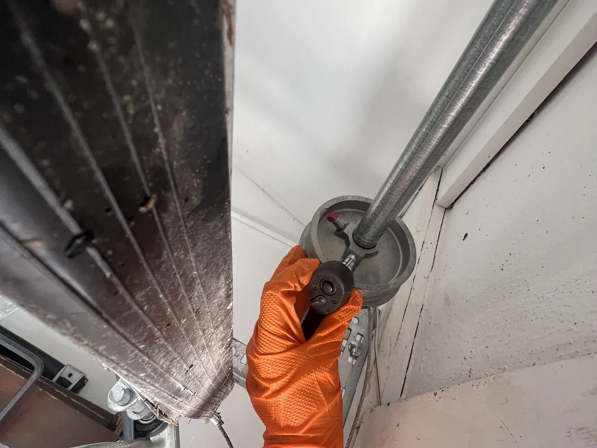 A gloved hand servicing the cable drum and bearing plate at the end of a garage door torsion shaft