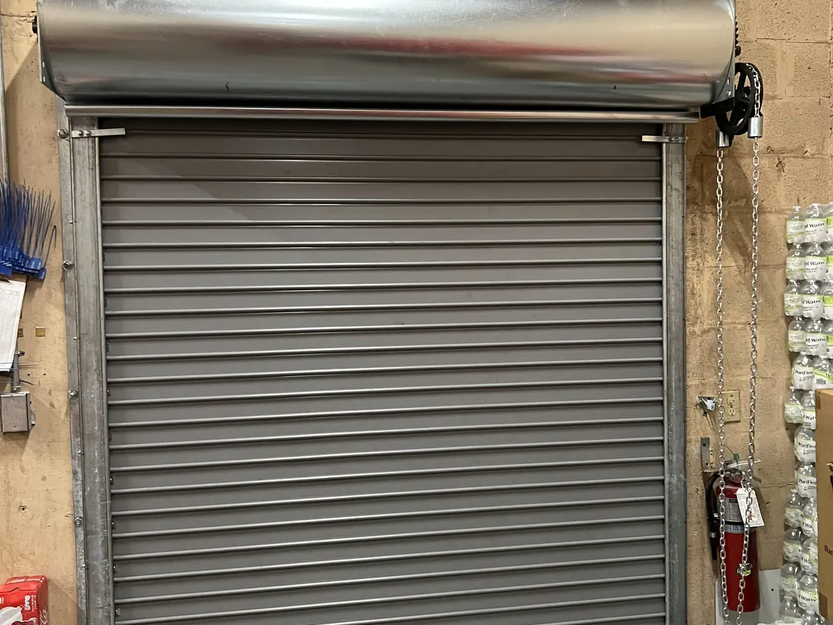 Grey coiling steel door closed in its guides beneath a galvanized hood, with the operating chain hanging beside it in a stockroom