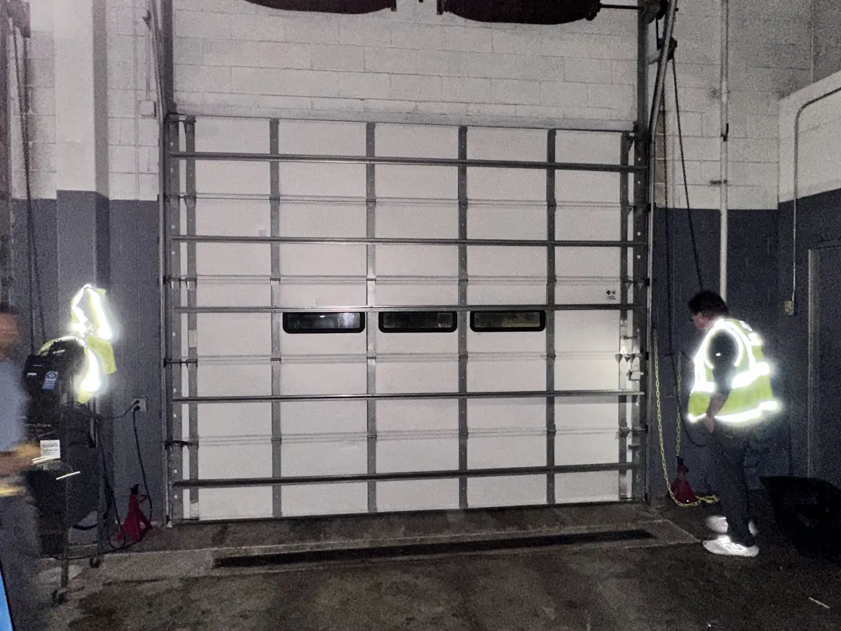 Technicians working on a white commercial sectional door with a full row of window panels, seen from inside the bay