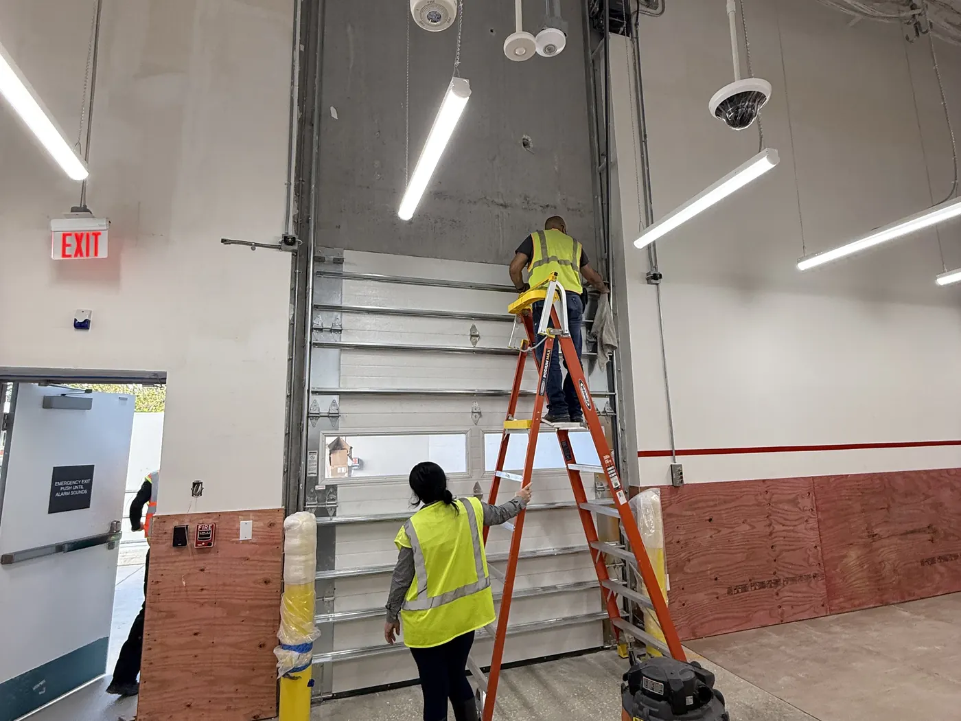 Two CMC technicians in high visibility vests fitting a sectional door in a commercial unit, one working from a ladder at the top section and one steadying the panel below
