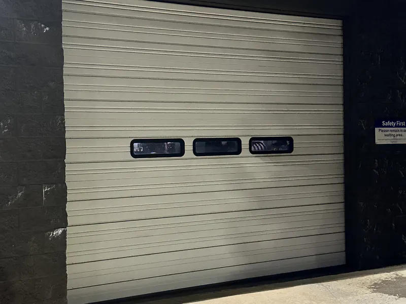 Beige steel commercial overhead door with three vision windows set in a dark block wall, photographed at night