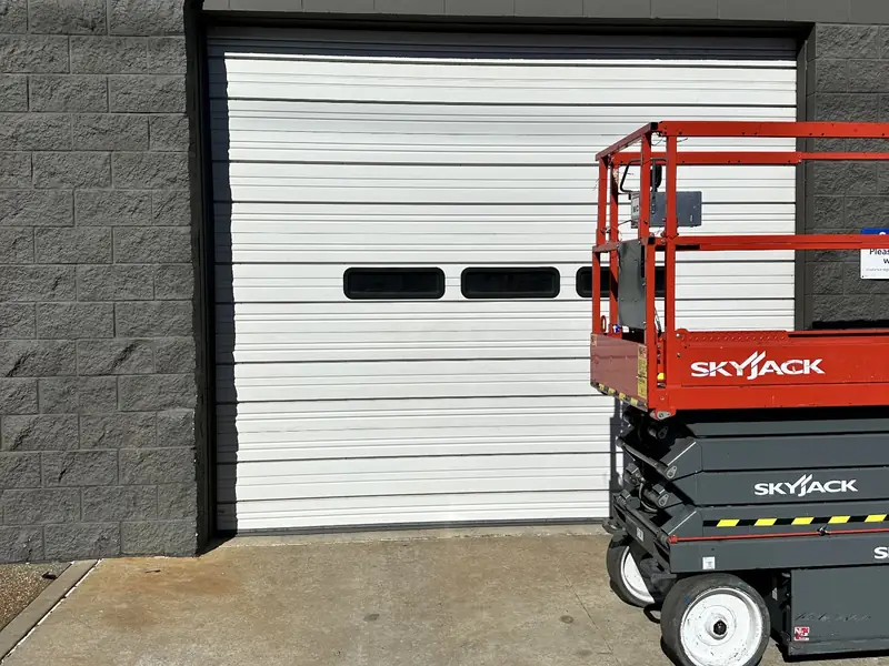 White steel commercial overhead door with a row of three vision windows set in a grey block wall, with a scissor lift parked alongside