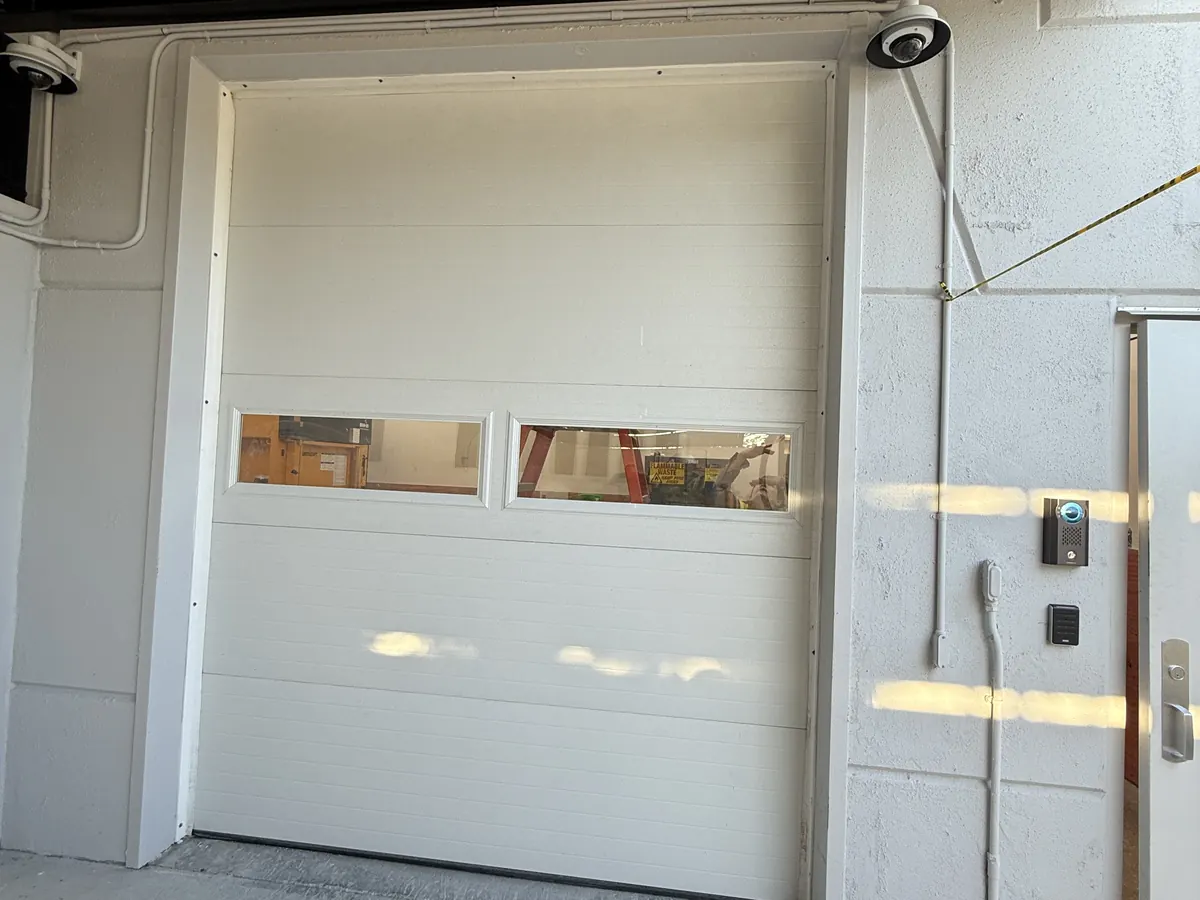 White sectional commercial door with two vision panels, closed in a concrete block wall beneath a dark canopy