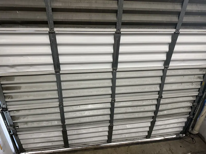 Interior of a closed white sectional garage door with galvanized horizontal struts and dark vertical reinforcement posts across the panels