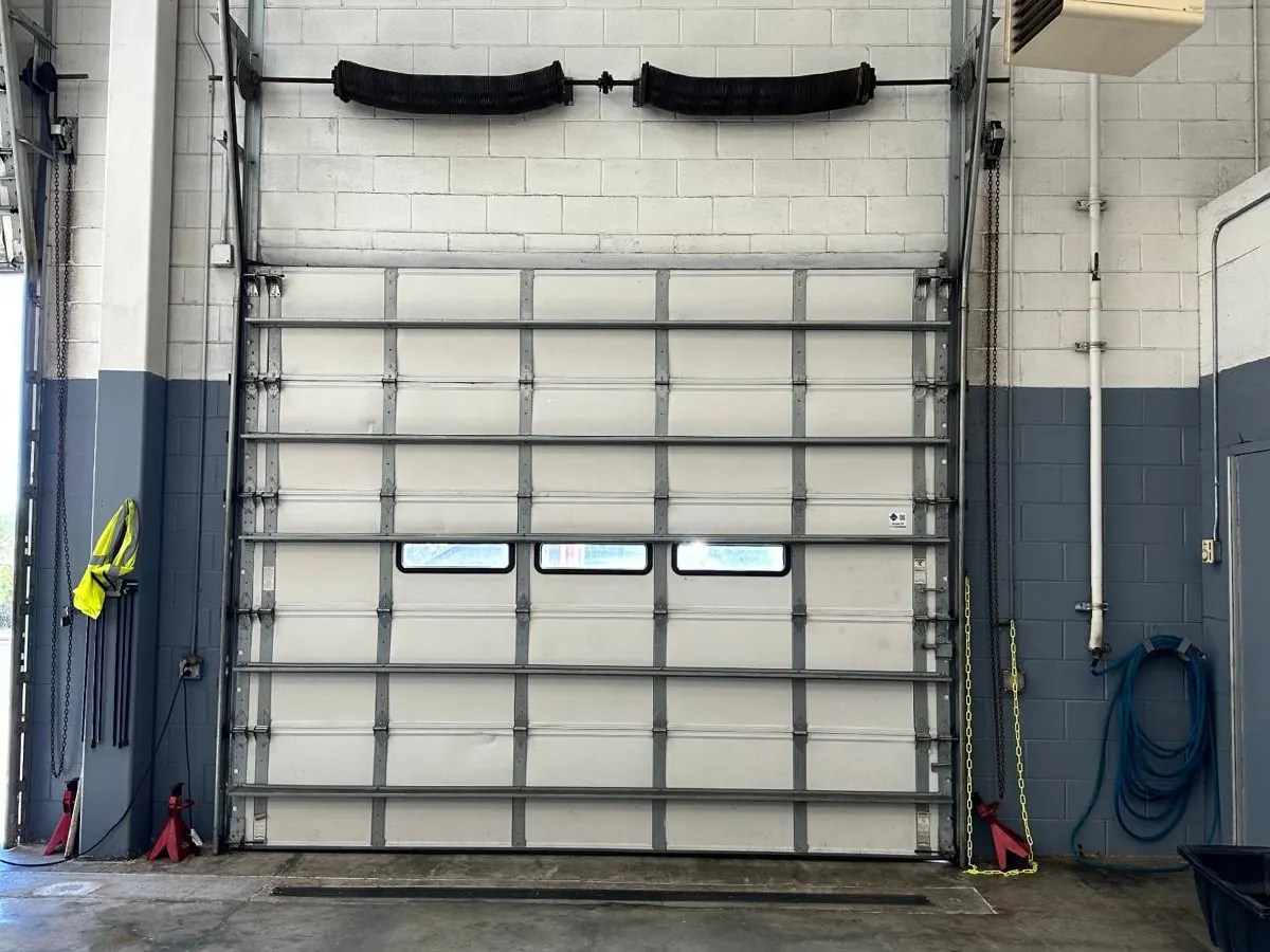 White insulated commercial sectional door with a row of vision panels, closed inside a service bay with the torsion springs visible above the opening