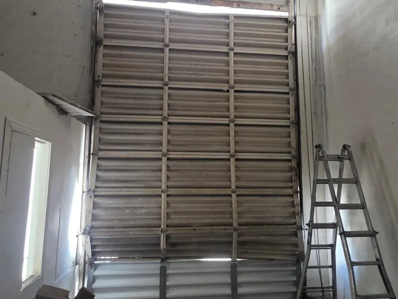 A worn corrugated steel commercial door seen from inside a building, with a step ladder beside it
