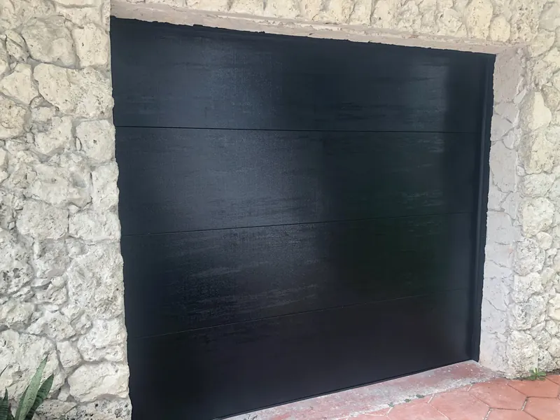 A flat black garage door with four horizontal sections set in a coral stone opening above terracotta tile