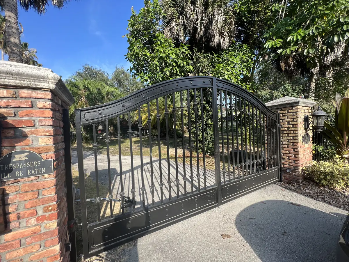 Arched ornamental iron double gate closed between brick pillars, the swing arm and its mounting fixed to the lower rail