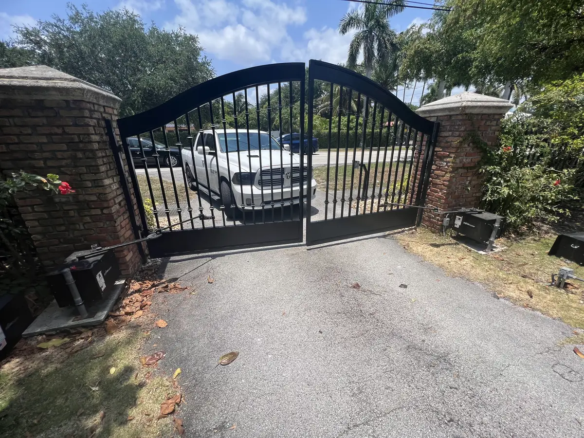 Arched ornamental iron double gate standing open between brick pillars, both swing operators visible at the base of the leaves