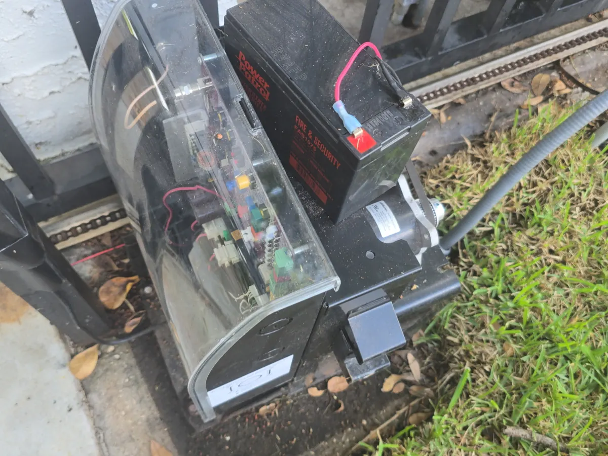 Gate operator housing set at the base of a metal gate, seen from ground level with the drive arm running to the leaf