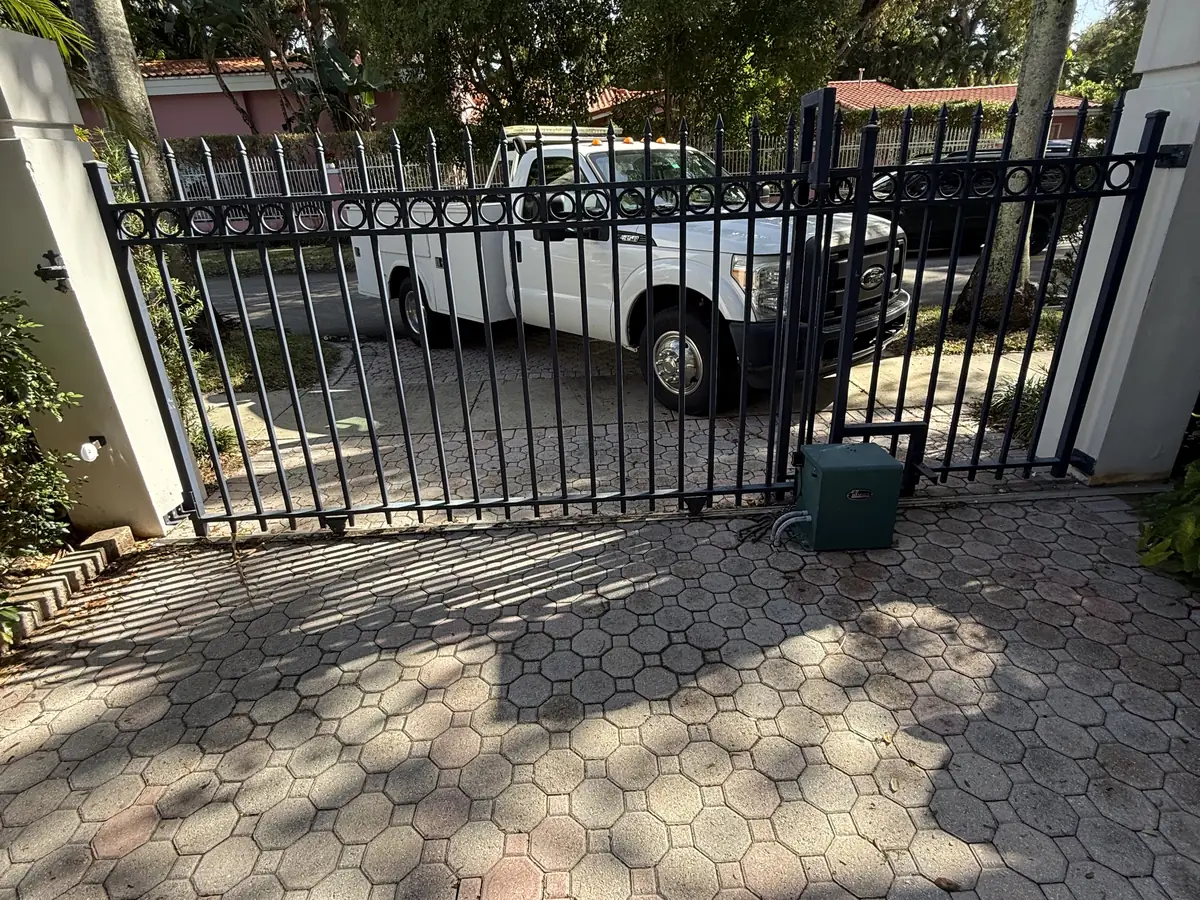 Ornamental iron swing gate standing open across a hexagonal paver driveway, the swing operator mounted low on the near leaf