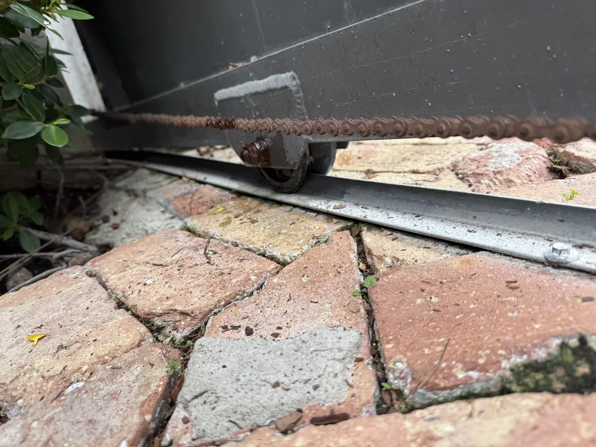 A rusted drive chain running along the bottom of a sliding gate, above the V groove wheel on its steel track