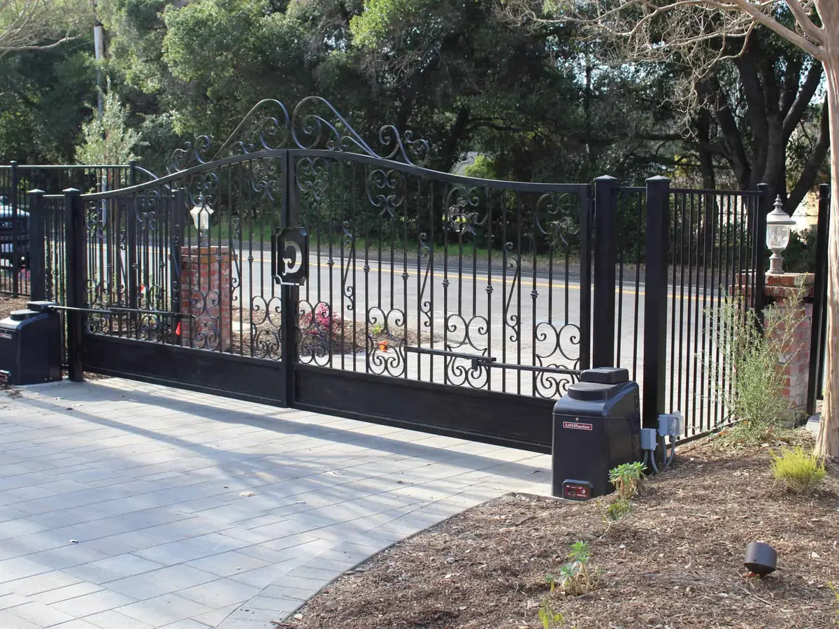 Black ornamental iron gate with a scrollwork arch top, closed across a wide residential driveway with the operator at the right hand post