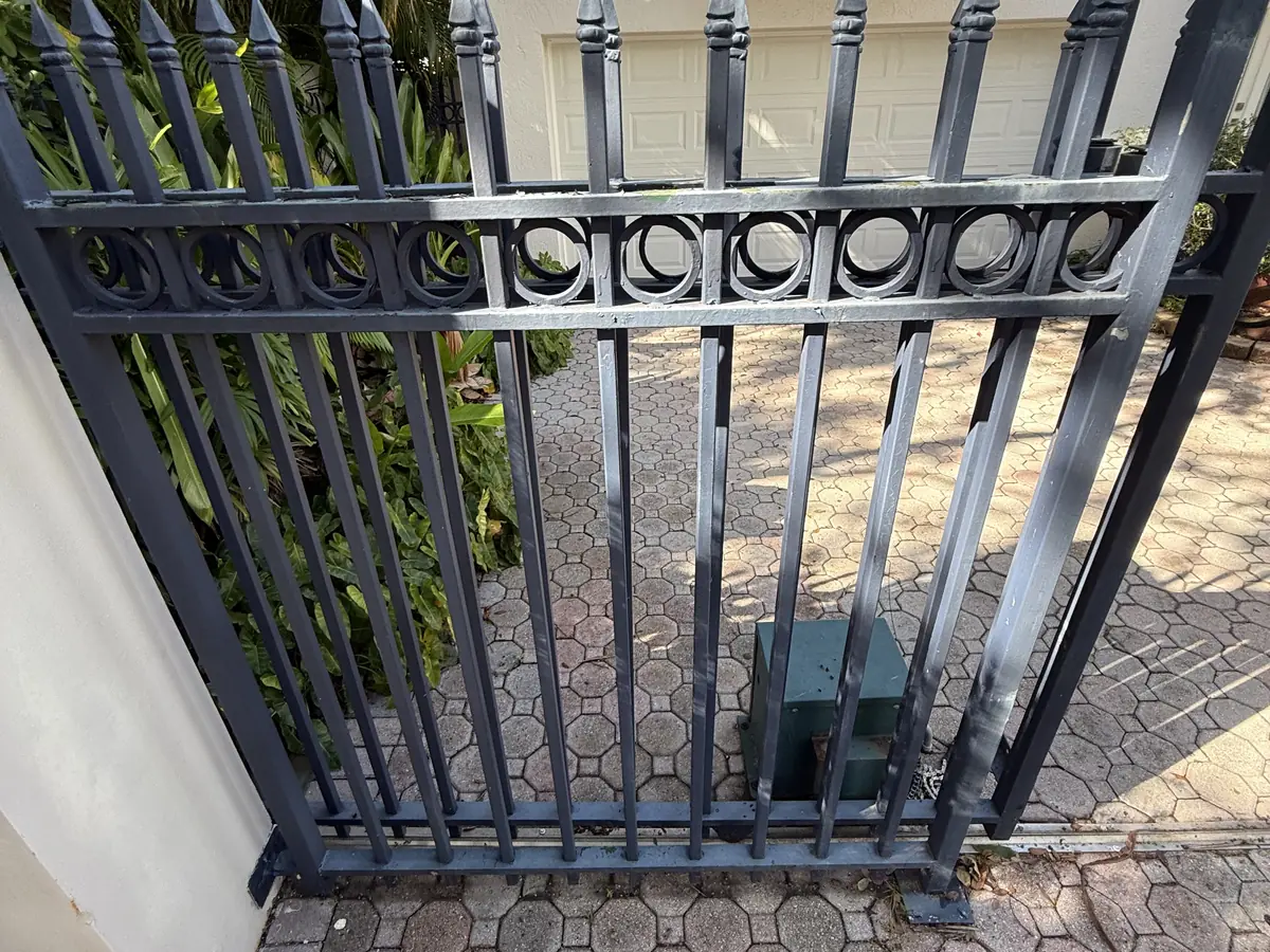 Spear top ornamental iron gate closed across a paver driveway, scrollwork running along the middle rail and the operator set at the base