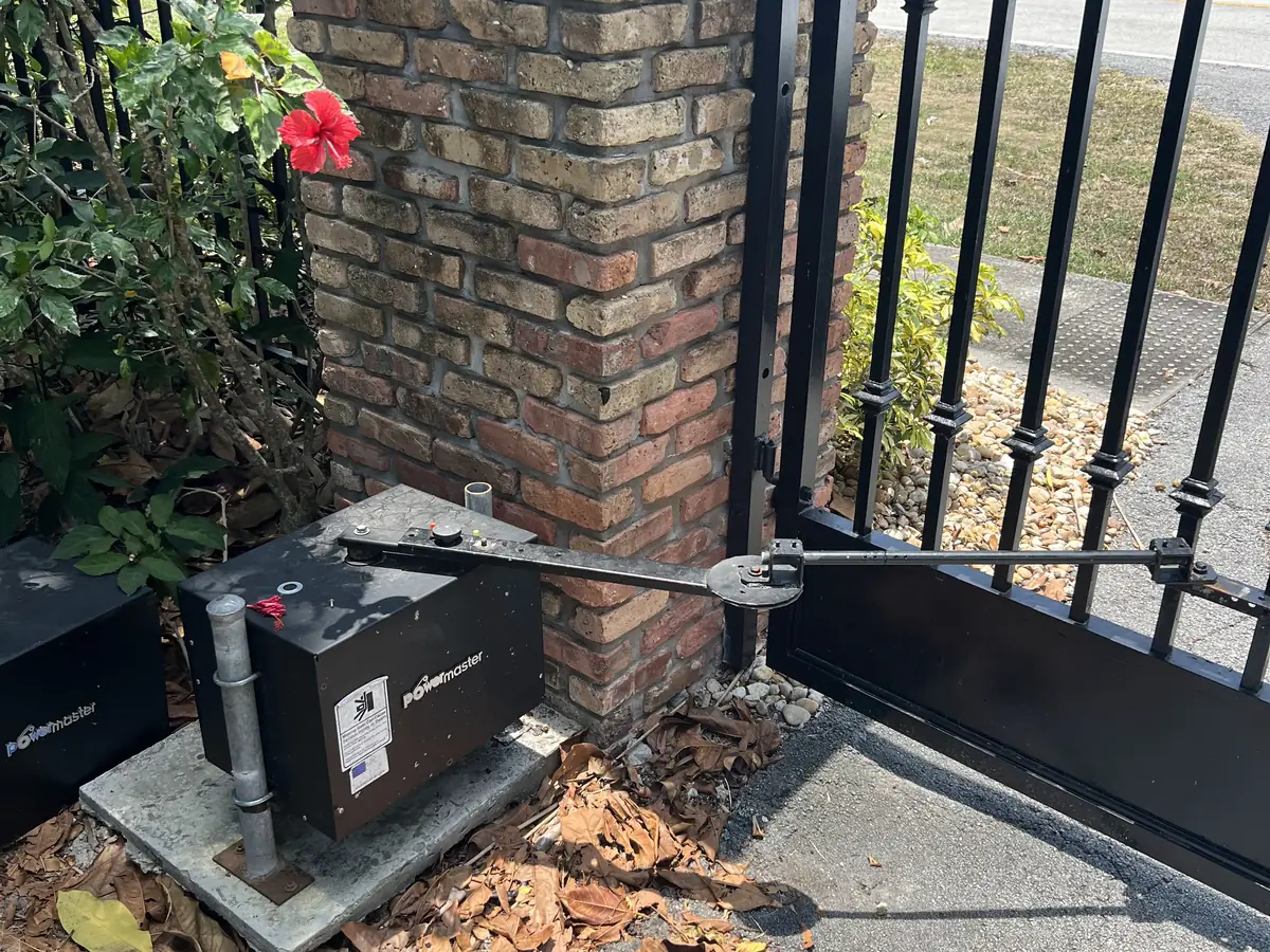 Swing gate operator and its arm mounted at a brick pillar, linked to the leaf of a black iron gate