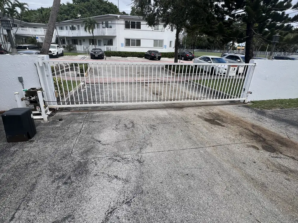 White tubular slide gate closed across the driveway of an apartment building, with the slide operator and its housing at the left hand post