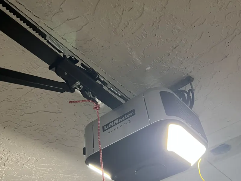 A LiftMaster garage door opener mounted on a garage ceiling with its belt rail and red emergency release cord attached