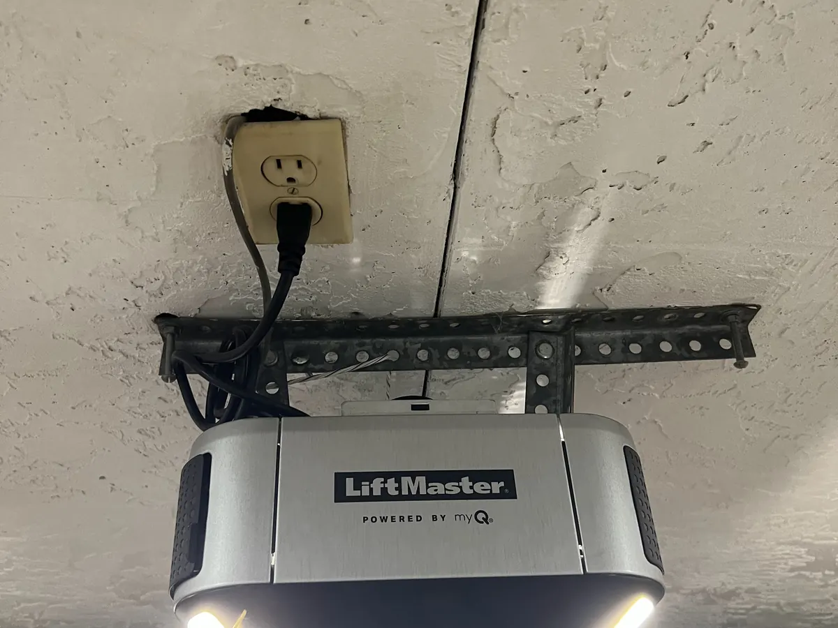 A silver and black LiftMaster garage door opener mounted to a garage ceiling with its lights on and its cord plugged into an outlet above