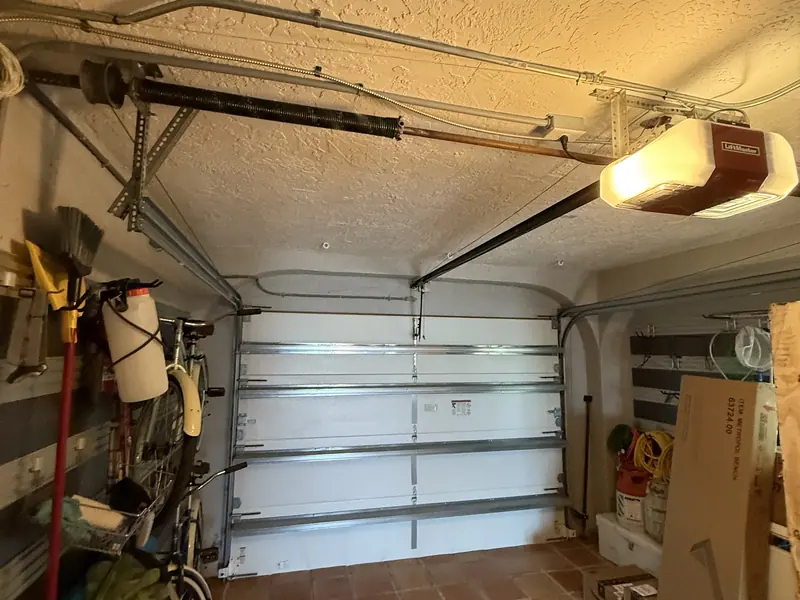 LiftMaster ceiling-mounted opener with its light on, its rail running to a closed white garage door, and the torsion spring mounted on an overhead shaft behind it
