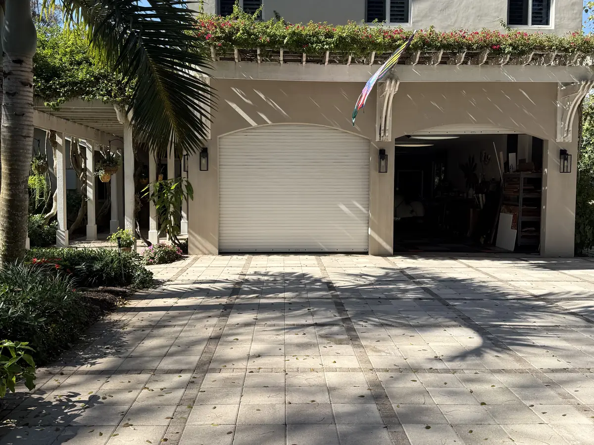 A white ribbed garage door under a first floor terrace, with a paver driveway and mature palms