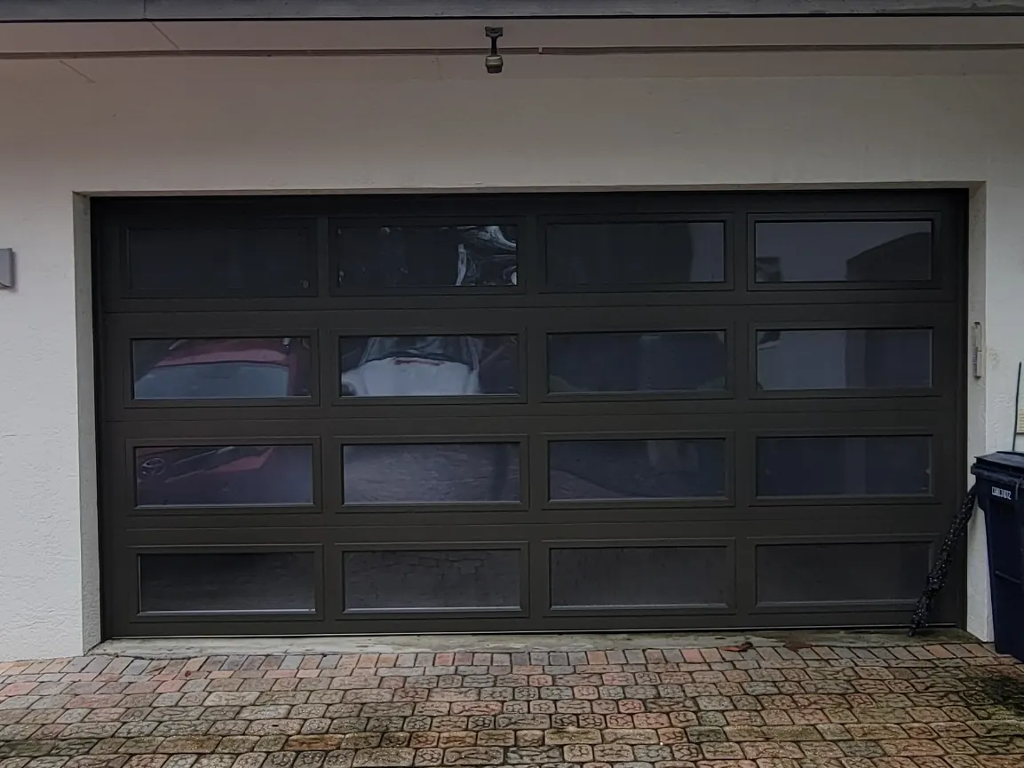 Dark modern flush double car garage door with a window row, above a cobblestone paver driveway