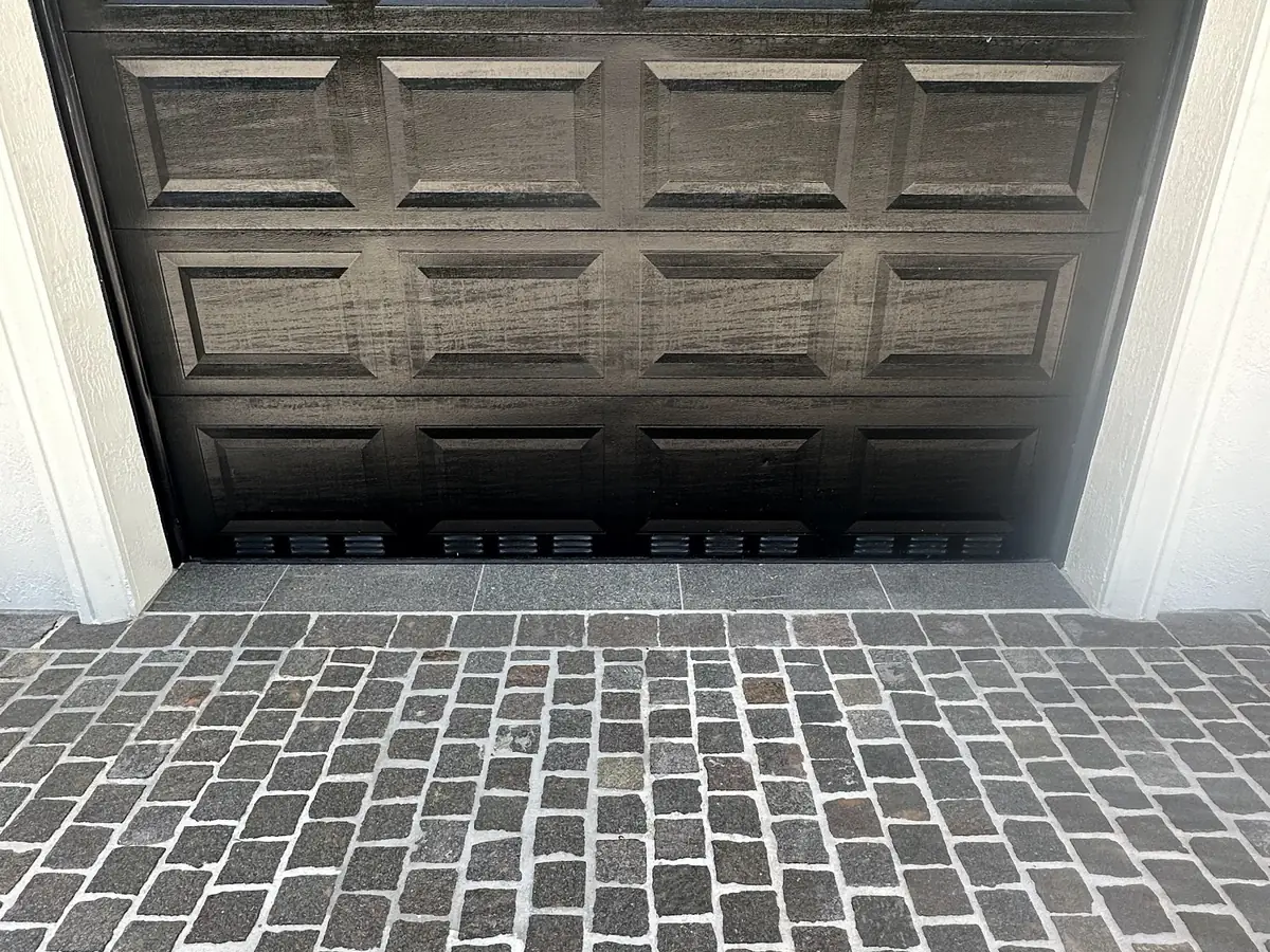 Black woodgrain raised panel garage door with ventilation slots, seen from inside the garage looking out over a cobblestone driveway