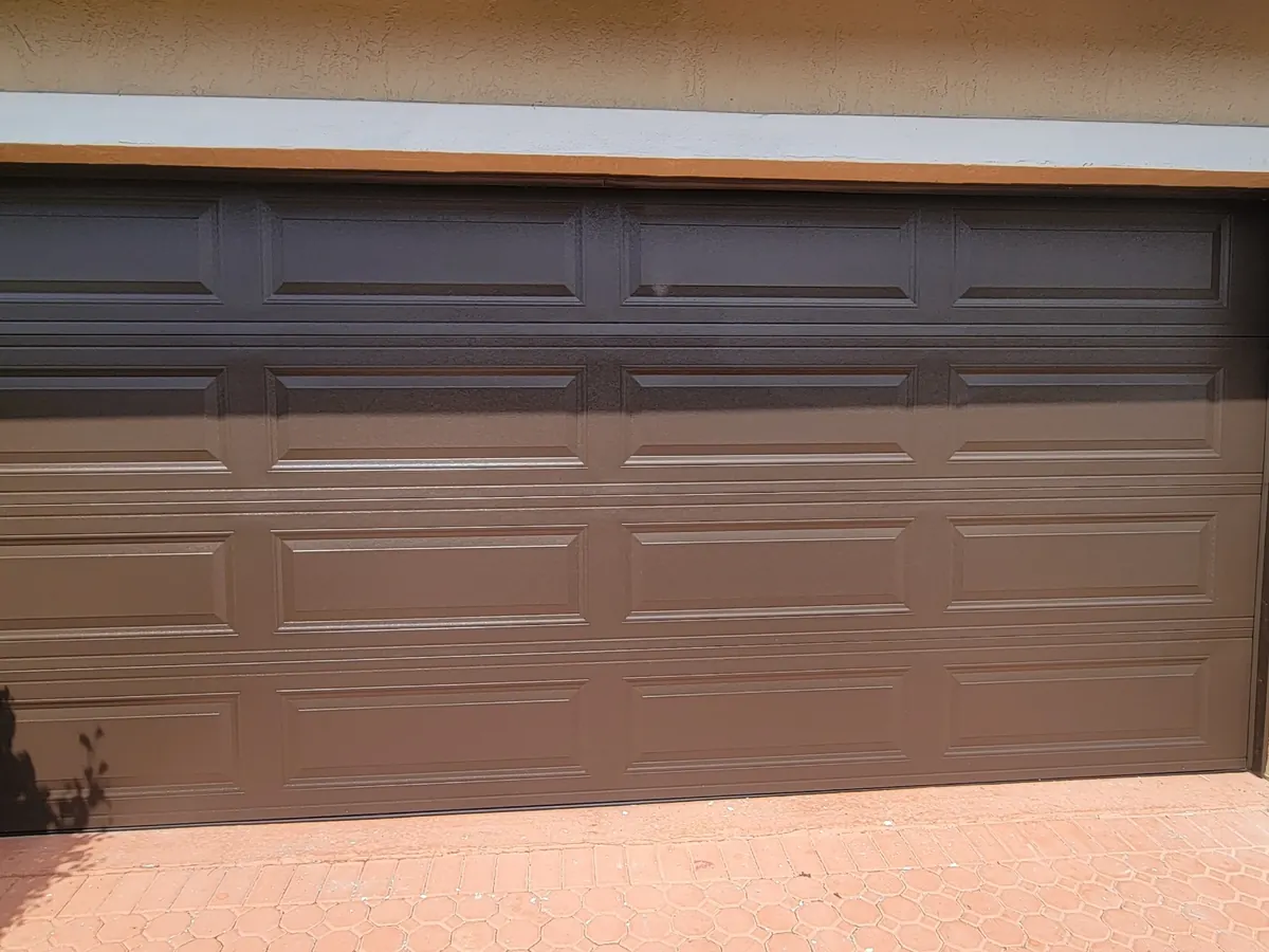 Bronze short panel double car garage door closed on a stucco home, with planting along the driveway edge