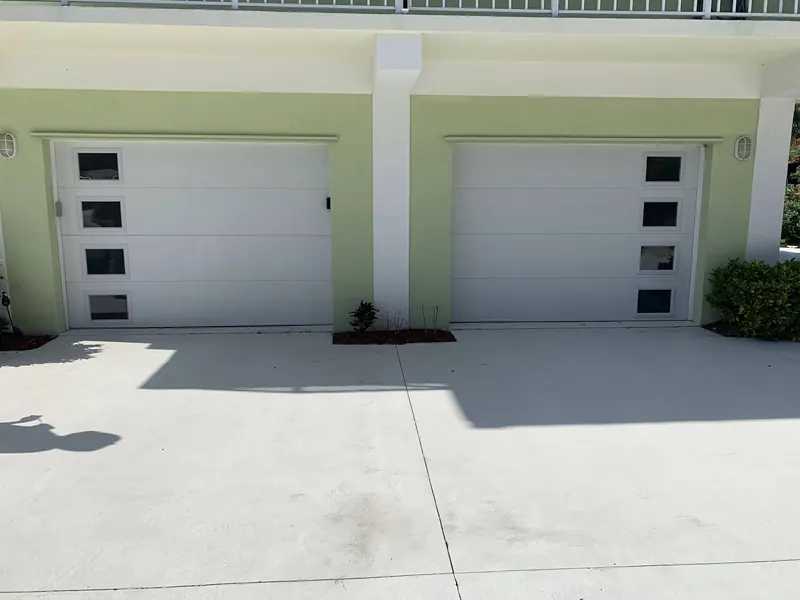 Two white single garage doors with square window rows on a green home
