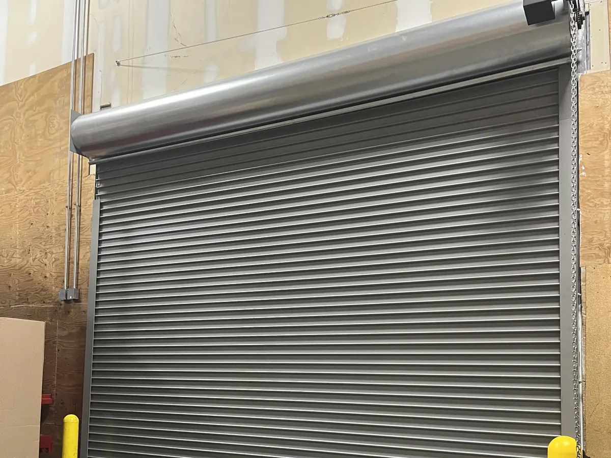 Grey rolling steel door closed inside a warehouse, with a galvanised metal hood over the coil above it and a hand chain running down the right side