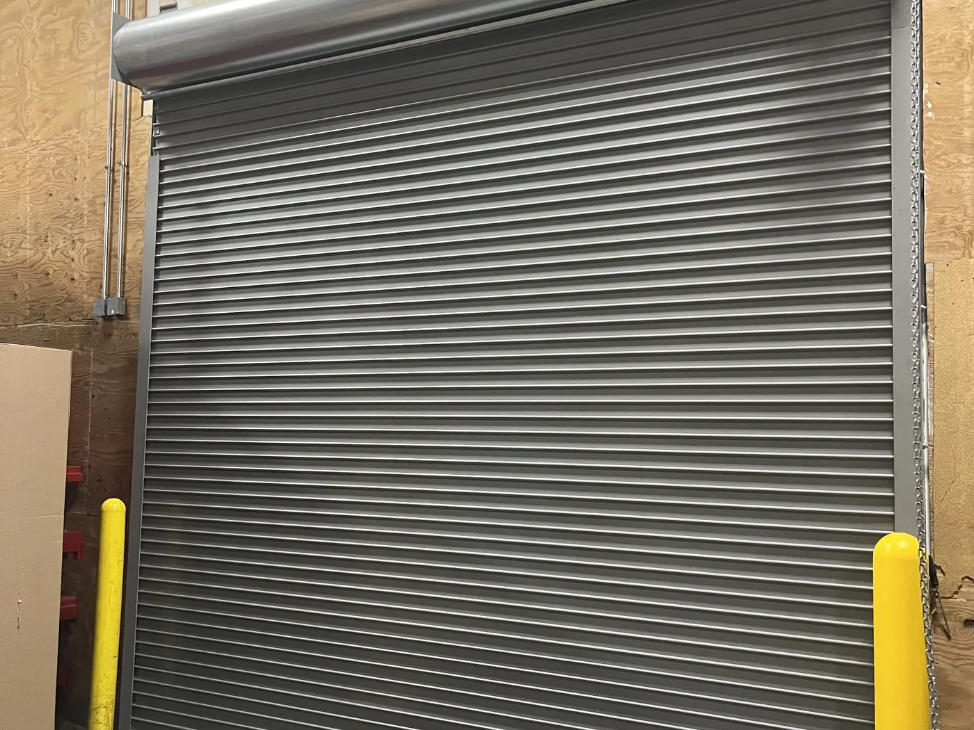A grey rolling steel door closed to the floor with the coil left exposed above it and bollards either side