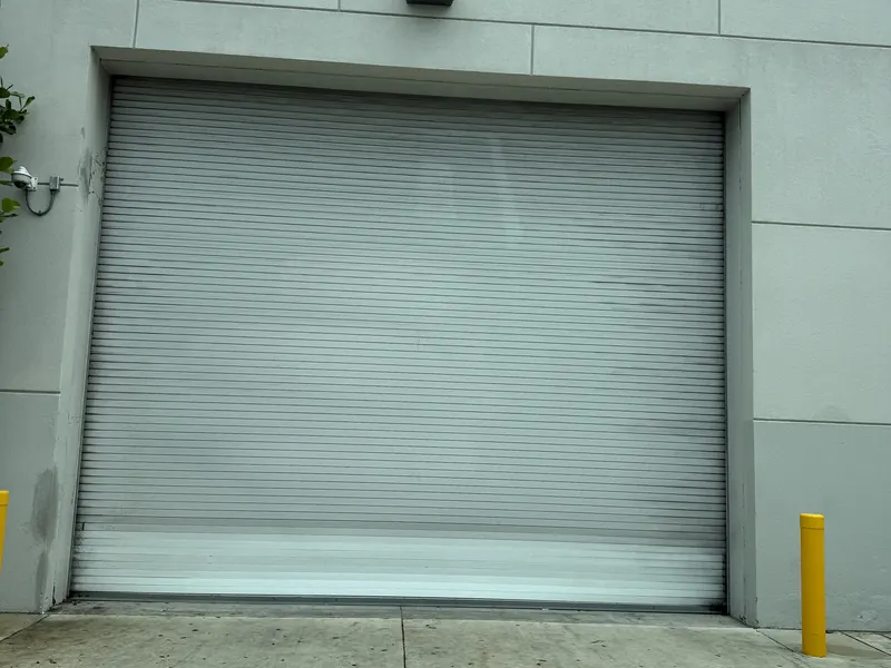 Grey coiling steel service door closed in the wall of a commercial building, with a yellow bollard at each side of the opening