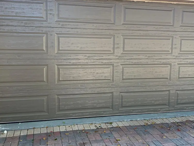 A taupe long-panel steel garage door closed in a stucco opening above a brick paver driveway
