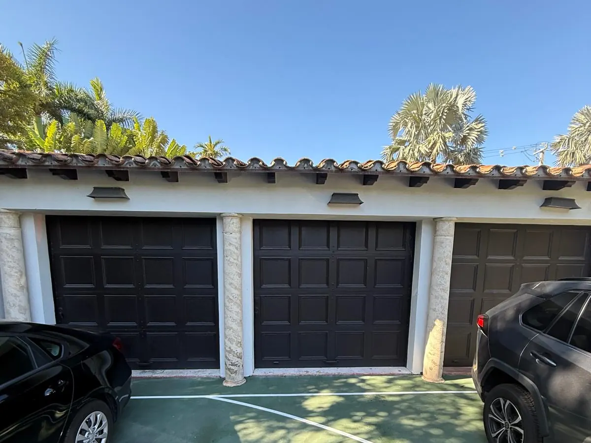 Three dark raised-panel garage doors on a tile-roofed building with cars parked in front