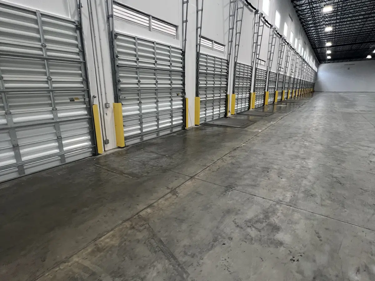 A row of closed commercial doors with yellow guard posts running down one wall of an empty warehouse