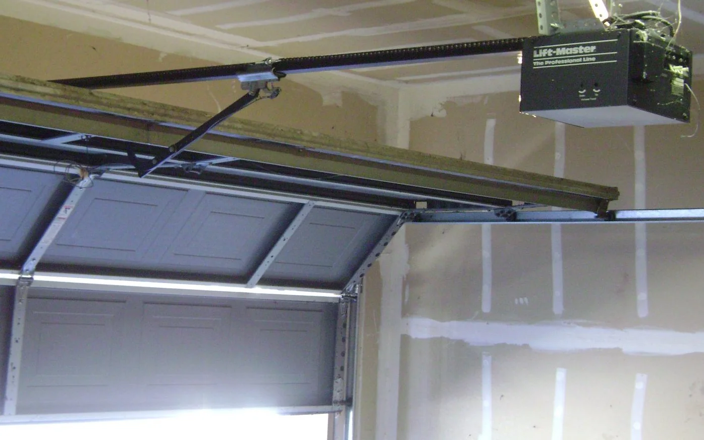 Inside a garage looking up at the opener: the motor unit on the ceiling, the rail running to the wall, the trolley arm connected to the top of the door, and the horizontal track