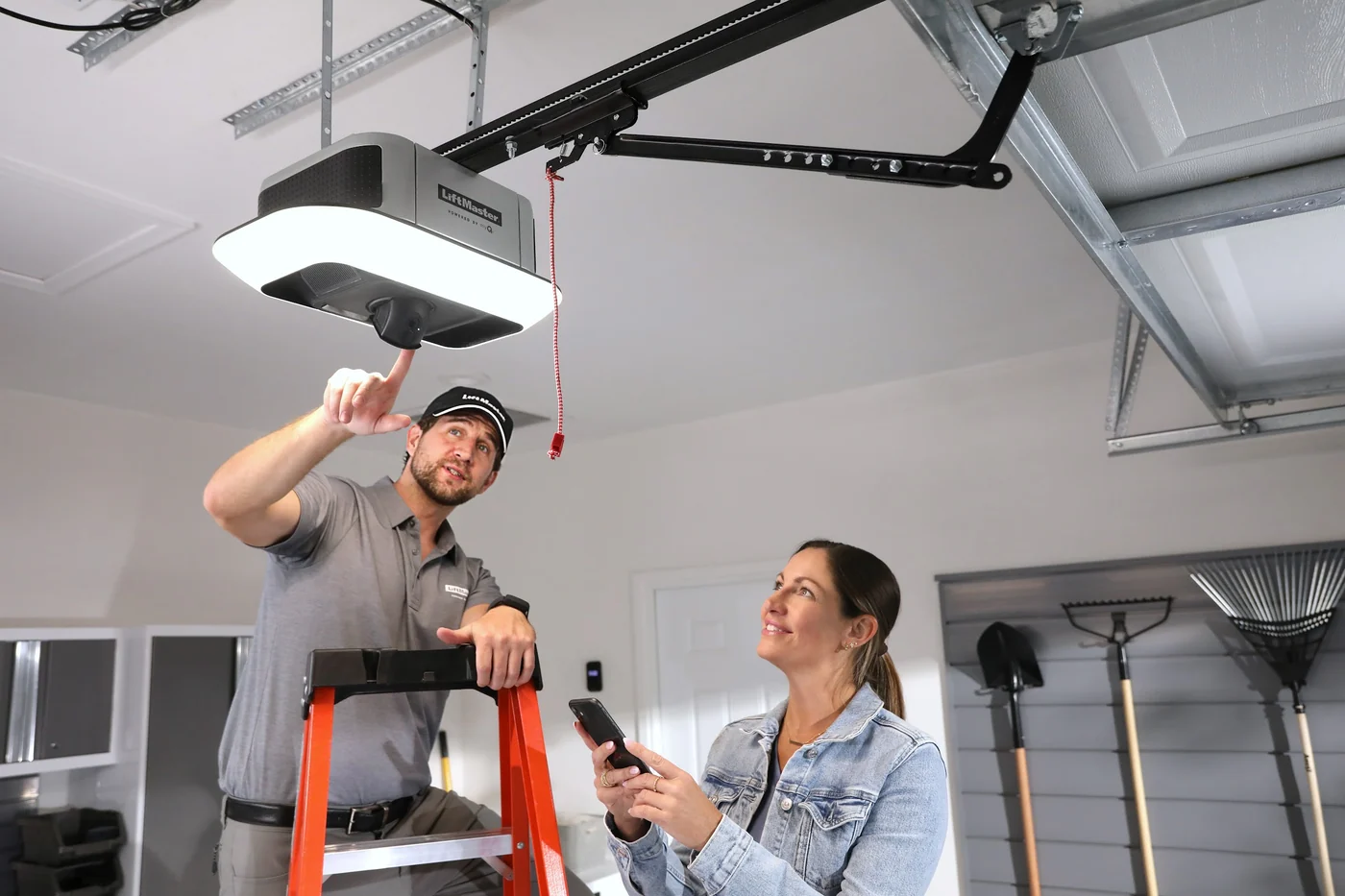 Technician on a ladder setting up a new LiftMaster opener while the homeowner pairs it to her phone