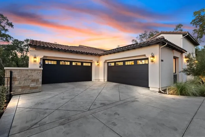 A Mediterranean style home at dusk with two dark garage doors and lit sconces